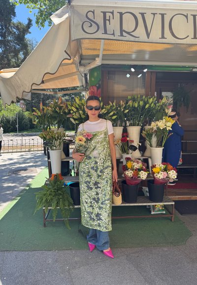 Vestido floral verde transparente con detalles rosas, llevado sobre una camiseta blanca. Sosteniendo un ramo, con zapatos de punta rosas y jeans de mezclilla visibles.
