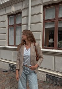 Ανοιχτό μπεζ σακάκι από λινό πάνω από λευκό πουκάμισο, συνδυασμένο με ξεθωριασμένα μπλε τζιν. Καφέ δερμάτινη τσάντα ώμου. Επίχρισμα από τούβλα και παράθυρο στο φόντο.