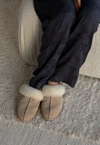 Bež copati iz semiša z mehko kremno kožuhasto manšeto. Ravna zasnova, rahlo zaokrožen prst in vidni šivi.