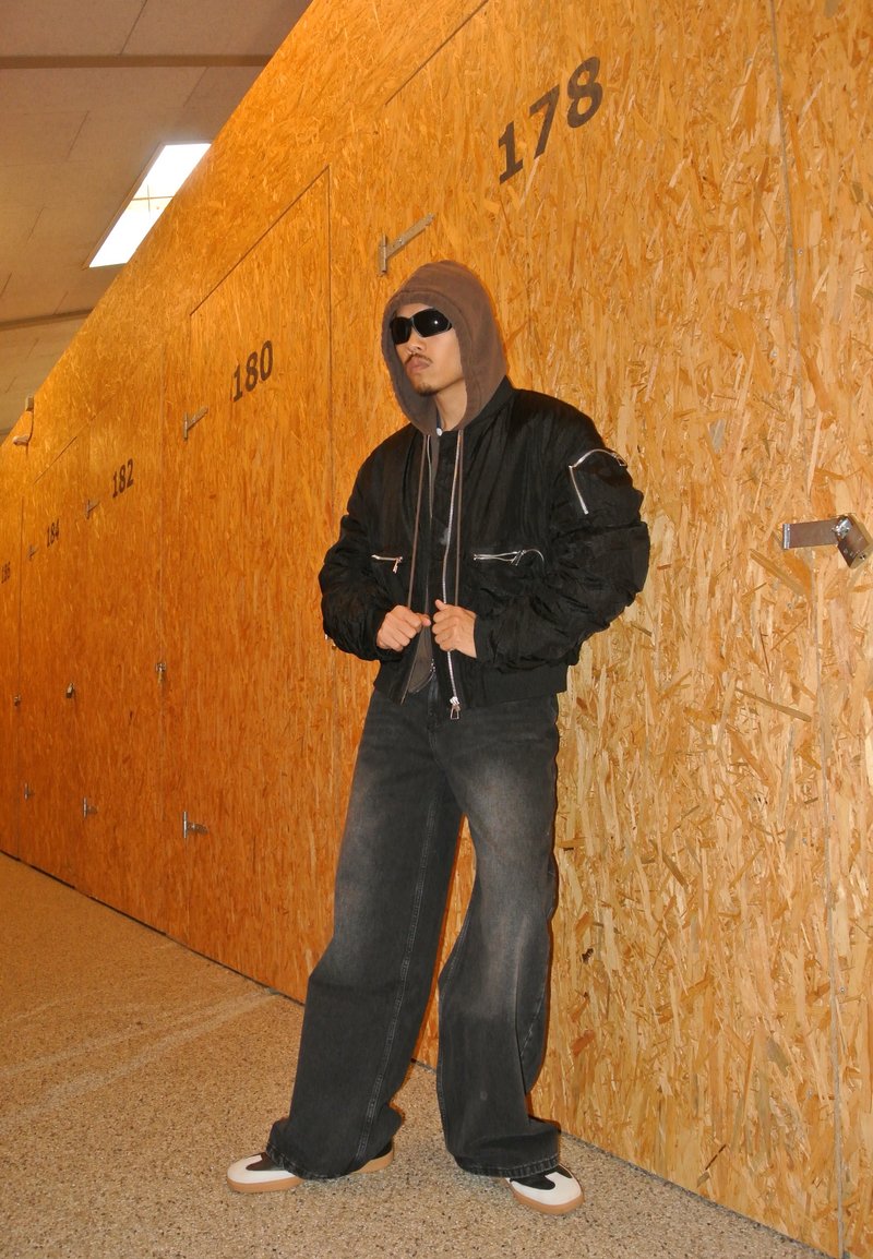 Μαύρο μπουφάν bomber με ασημένιους φερμουάρ, γκρι φούτερ, φαρδύ τζιν παντελόνι σε σκούρο χρώμα και μαύρα αθλητικά παπούτσια με μπεζ λεπτομέρειες, σε φόντο πορτοκαλί τοίχο.