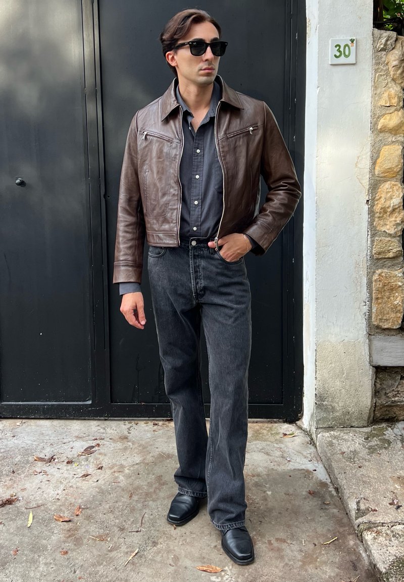 Jaqueta cropped de couro castanho com zíperes, camisa cinza de botões, jeans pretos e sapatos pretos. Aposentado contra uma parede escura.