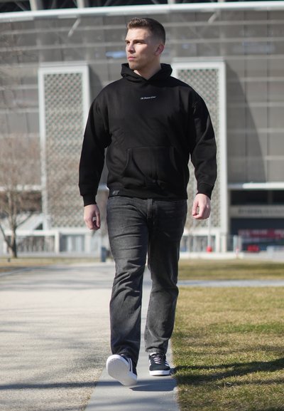 Hombre con sudadera con capucha negra, jeans grises y zapatillas negras caminando por un sendero pavimentado en una zona urbana al aire libre durante el día.