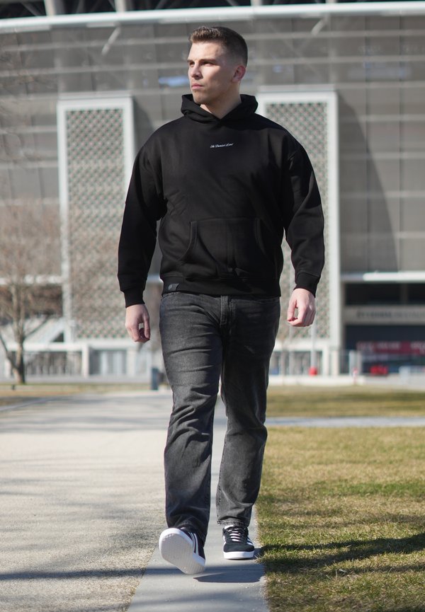 Bărbat purtând hanorac negru, blugi gri și adidași negri, mergând pe o alee pavată într-o zonă urbană în aer liber, în timpul zilei.