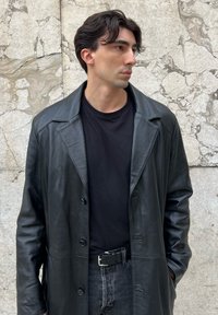 Casaco trench de couro preto com colarinho notched e fechos de botão, combinado com uma camisa preta e jeans escuros contra uma parede texturizada.
