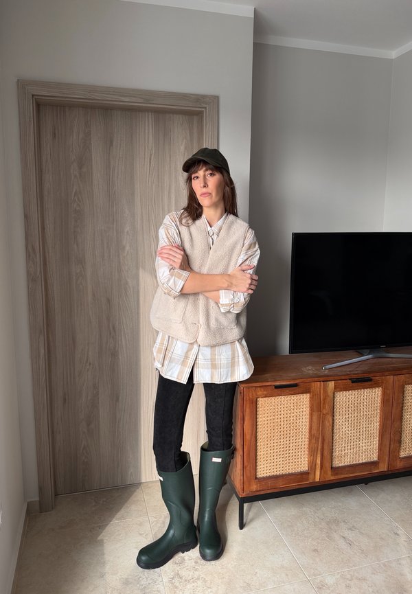 Frau in beigefarbener Weste, kariertem Hemd, schwarzer Hose, grünen Gummistiefeln und dunkler Kappe steht drinnen neben einer Holztür und einem TV-Regal.