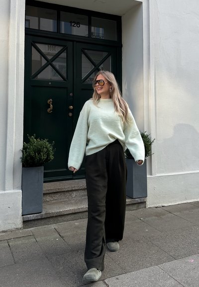 Suéter oversized verde menta con puños acanalados, pantalones anchos negros y calzado gris. Puerta verde oscura y plantas en macetas en el fondo.
