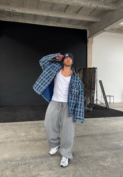 Pantalones de chándal grises sueltos, una camiseta sin mangas blanca, una camisa de cuadros azul y negra de sobrecamisa, y un gorro oscuro con un logotipo. Zapatillas blancas y grises.