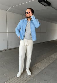 Chaqueta de denim azul claro sobre una camiseta gris, combinada con pantalones blancos y zapatillas blancas. El entorno presenta un fondo de concreto.