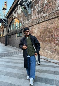 Homme souriant portant des lunettes, un béret noir, un long manteau noir, un gilet vert et un jean bleu, marchant dans une rue de la ville avec des murs en briques et des graffitis.