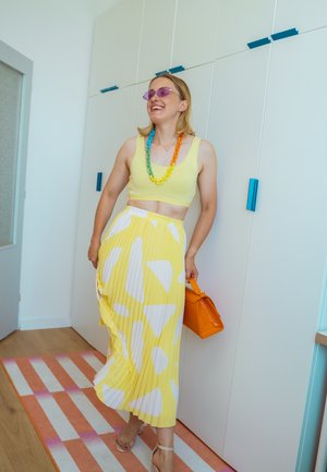 Falda amarilla con pliegues y formas blancas, combinada con un top corto amarillo. Los accesorios incluyen un collar multicolor y un bolso naranja.