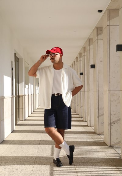 Camisa blanca de textura con manga corta, camiseta blanca, pantalones cortos de mezclilla oscuros, gorra roja, gafas, zapatos negros y calcetines blancos hasta el tobillo. Entorno de pasillo iluminado.