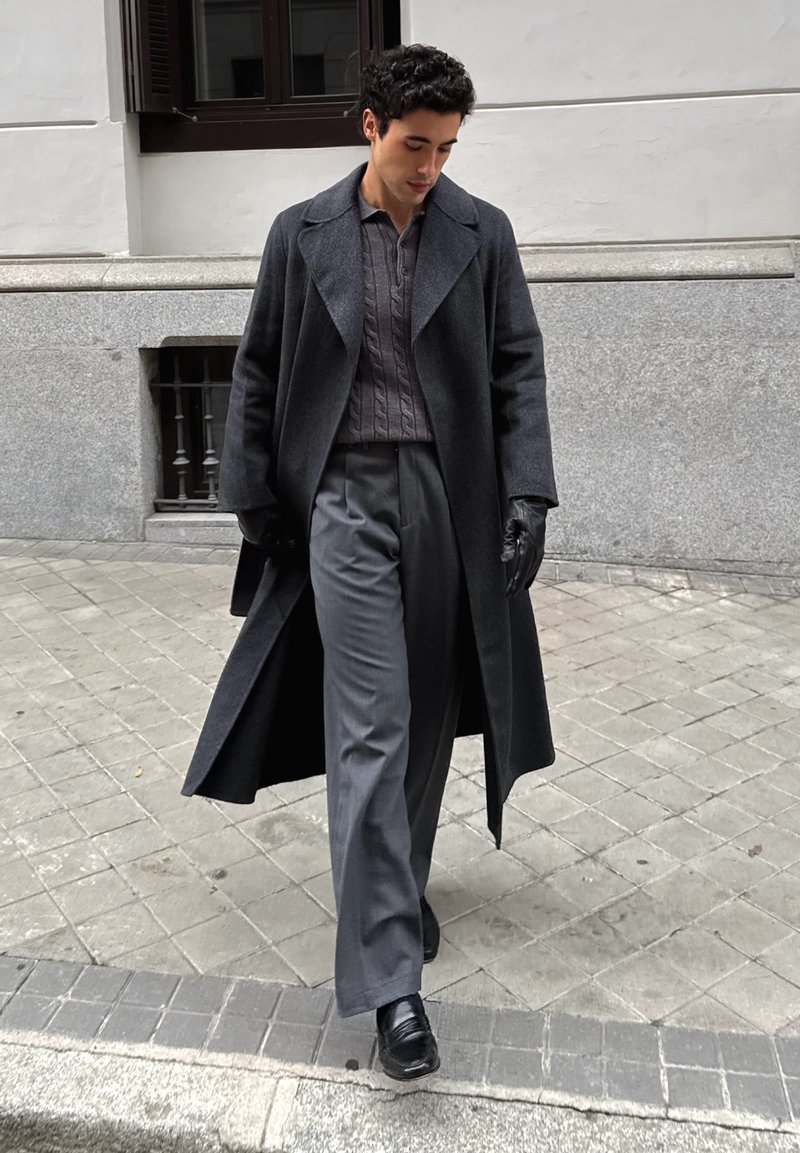 Homme marchant sur un trottoir en ville, portant un long manteau sombre, un pull gris à mailles torsadées, un pantalon large gris, des gants noirs et des chaussures noires.