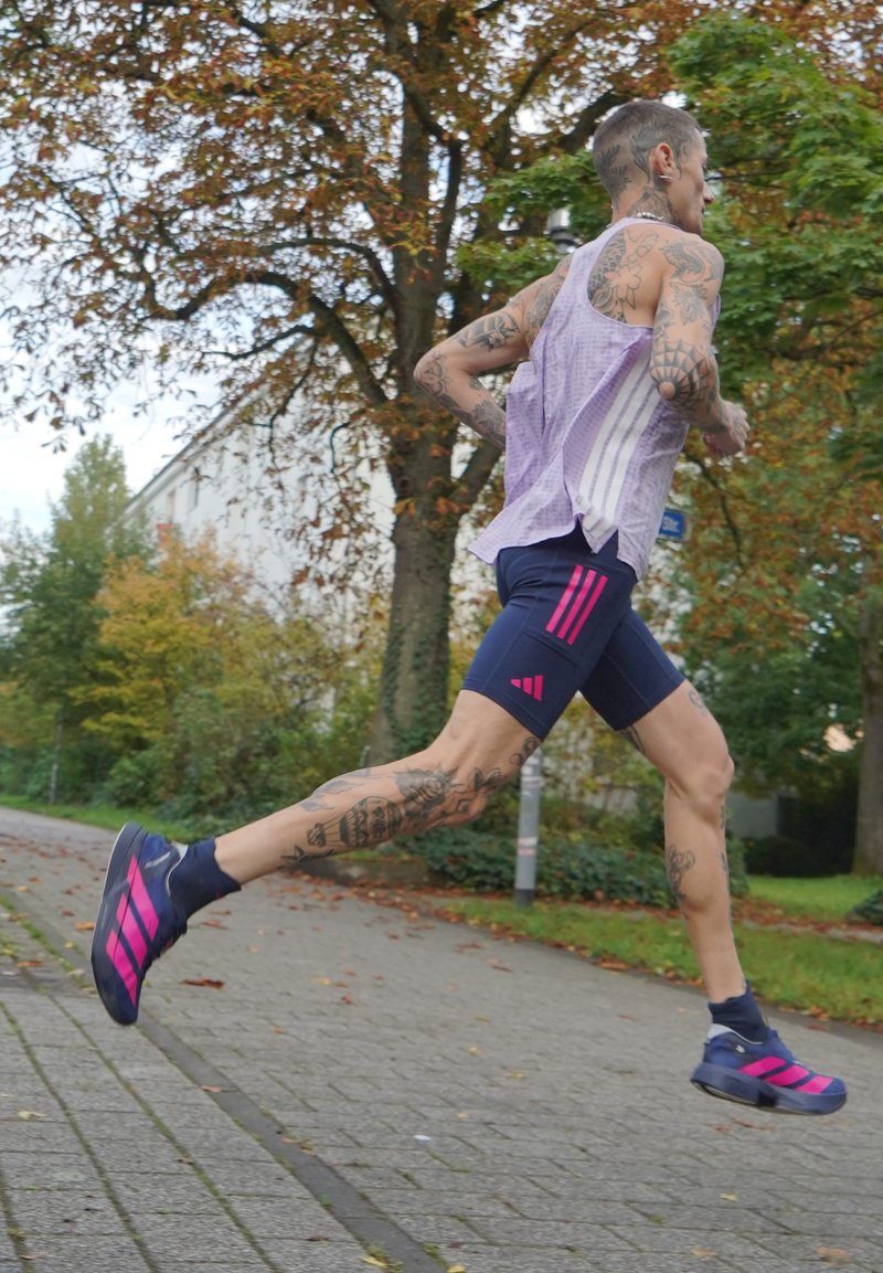 Corredor com tatuagens, vestindo uma t-shirt roxa e calções azul-marinho com riscas rosas, a correr ao ar livre num caminho pavimentado no outono.