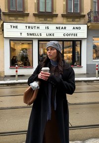 Jeune femme en manteau et chapeau tenant une tasse de café devant un café avec un panneau indiquant "La vérité est proche et elle sent le café."