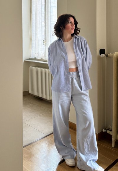 Camisa de lino azul claro con botones sobre una parte superior blanca corta, combinada con pantalones sueltos azul claro y zapatillas blancas, de pie en el interior.