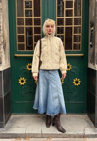 Gepolsterte cremefarbene Kapuzenjacke, hellblaue Jeans-A-Linien-Rock, braune High-Heels-Stiefel, stehend vor einer grünen Tür mit Sonnenblumenakzenten.