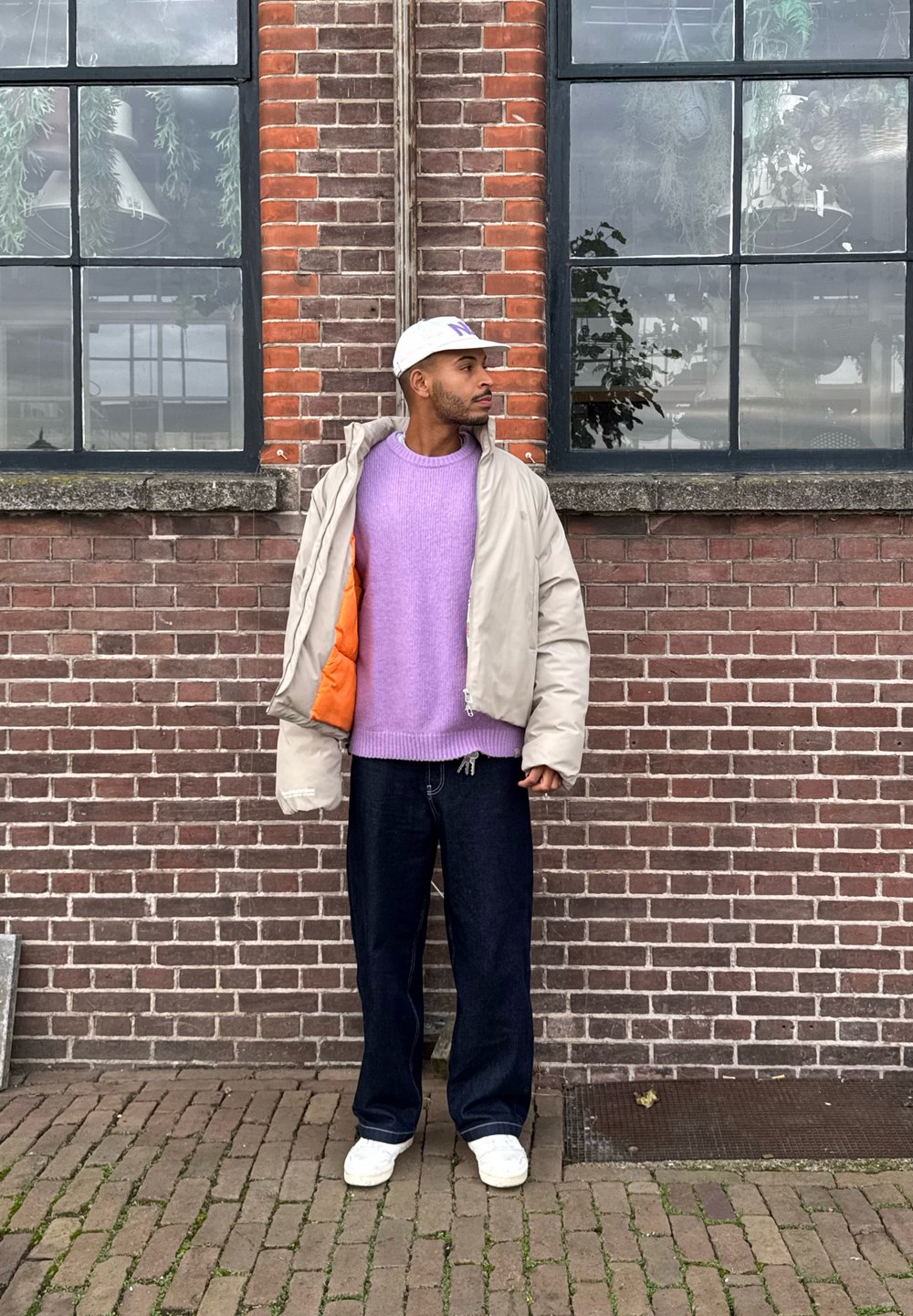 Gewatteerd jack in beige met een oranje voering, gedragen over een lichtpaarse trui. Donkere jeans en witte schoenen, tegen een achtergrond van een bakstenen muur.