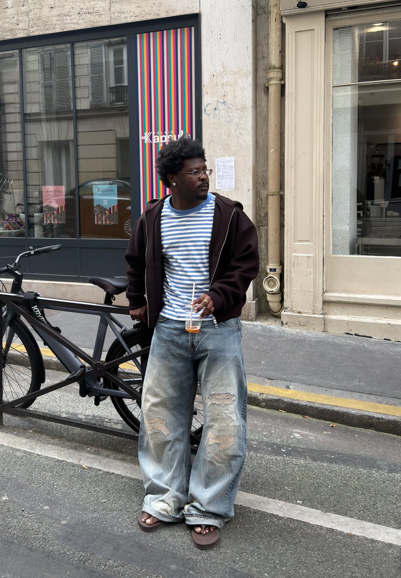 Man som bär en randig skjorta, mörk jacka och trasiga vida jeans står vid en svart cykel och håller en dryck på en stadsgata.