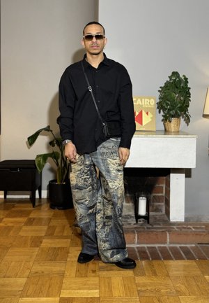 Hombre con camisa negra de gran tamaño, pantalones anchos con estampado, zapatos negros, gafas de sol y bolso cruzado, de pie en el interior junto a una chimenea y plantas.