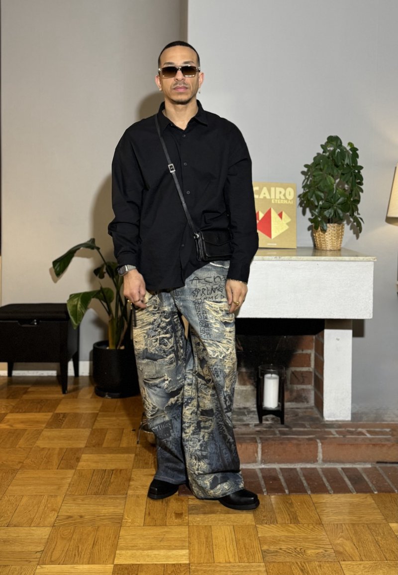 Homme portant une chemise noire surdimensionnée, un pantalon large à motifs, des chaussures noires, des lunettes de soleil et un sac en bandoulière, debout à l'intérieur près d'une cheminée et de plantes.