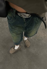 Pantalones cortos de mezclilla en un lavado oscuro con un ajuste relajado, combinados con crocs color beige y calcetines blancos. Un cinturón negro con una hebilla plateada completa el conjunto.
