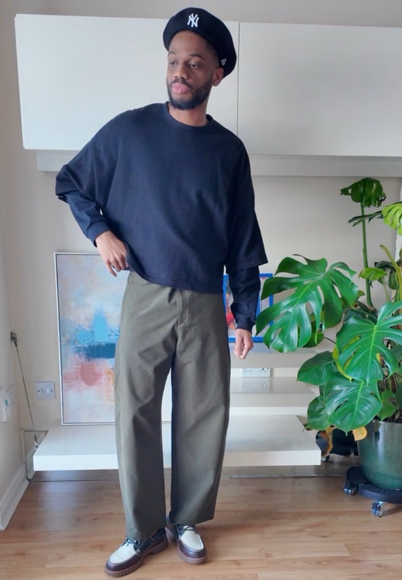 Black long-sleeve cropped sweatshirt, olive green wide-leg pants, and two-tone shoes with white upper and dark sole. Plant visible in background.