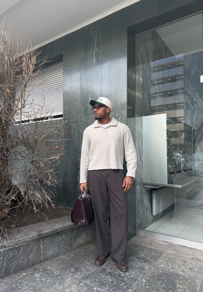 Hombre con suéter beige, gorra y gafas, de pie frente a un edificio moderno sosteniendo una bolsa marrón oscuro junto a un arbusto sin hojas.