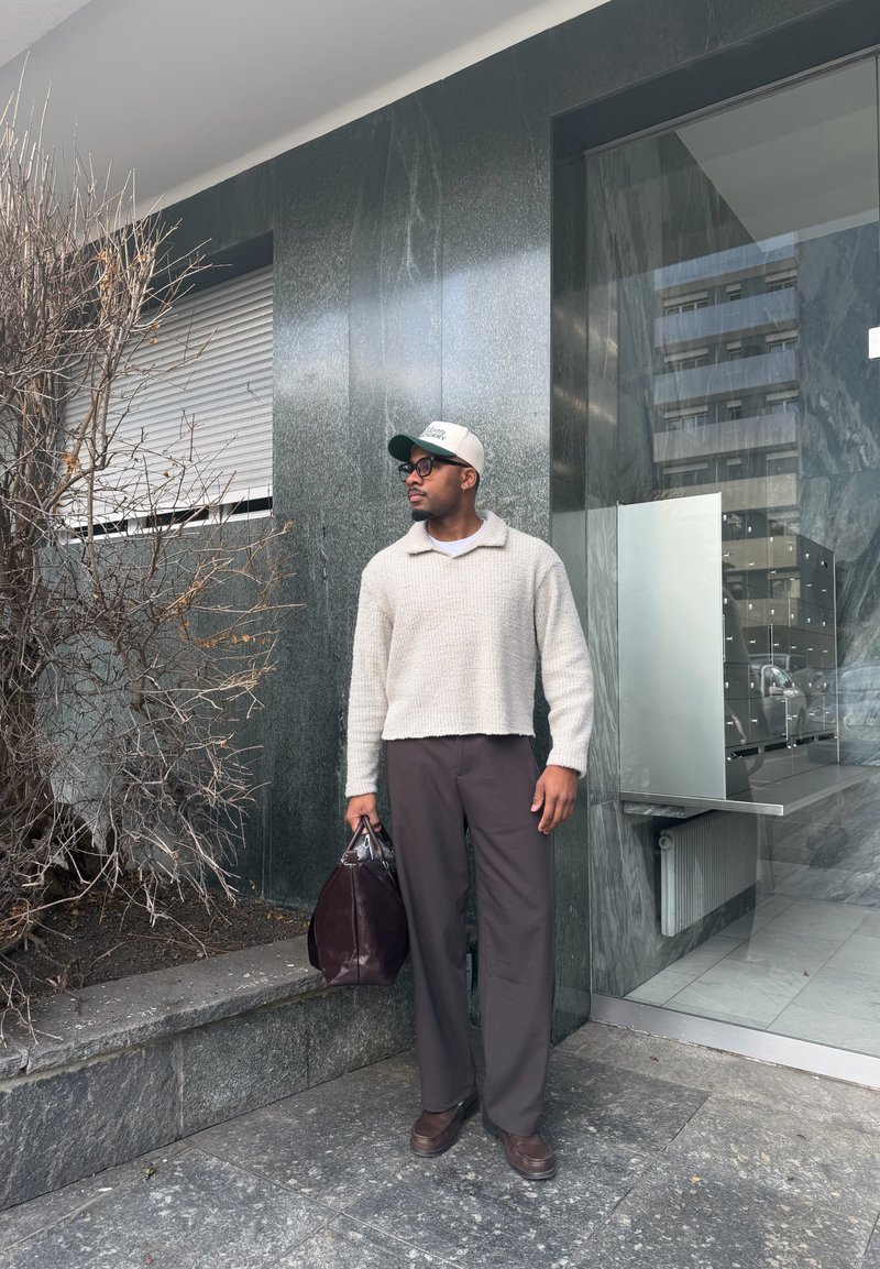 Homme portant un pull beige, une casquette et des lunettes, debout devant un bâtiment moderne, tenant un sac brun foncé près d'un arbuste nu.