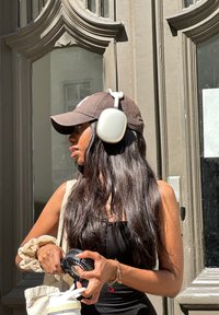 Ασύρματα ακουστικά σε λευκό χρώμα, φορεμένα πάνω από ένα σκούρο καπέλο του μπέιζμπολ, με μακριά σκούρα μαλλιά, κρατώντας έναν φορητό ανεμιστήρα και μια ελαφριά τσάντα.