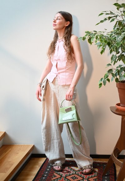 Blusa sin mangas rosa con la parte delantera anudada, pantalones de pierna ancha beige y un bolso estructurado verde claro. Fondo texturizado con plantas y escaleras.