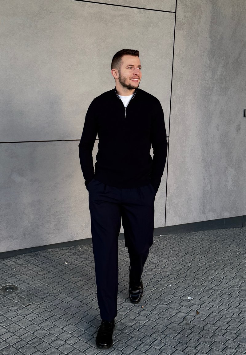 Young man walking on a paved pavement, hands in pockets, wearing a black zip-up jumper, dark trousers, and black shoes, smiling and looking to the right.