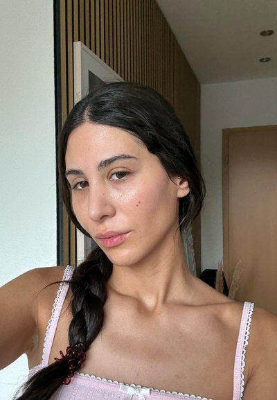 Retrato de una persona con cabello largo y oscuro, peinado en una trenza, que lleva una camiseta rosa clara con encaje, ubicado frente a un fondo neutro.