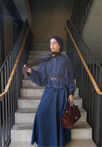 Cardigan tricoté bleu marine avec un motif texturé, associé à une longue jupe en denim. Ceinturon marron, sac à main bordeaux et écharpe foncée complètent la tenue.