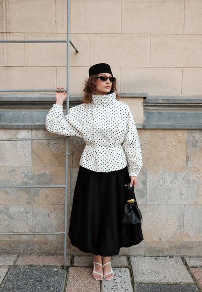 Mujer con sombrero negro, gafas de sol, chaqueta blanca con lunares, falda negra y sandalias blancas que sostiene un bolso negro, de pie junto a una escalera metálica en una pared de piedra.