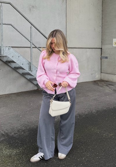 Mujer rubia con suéter rosa, pantalones grises de pierna ancha y zapatillas blancas sosteniendo un bolso blanco y gafas de sol junto a una escalera de concreto.