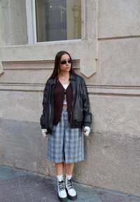 Μαύρο δερμάτινο μπουφάν, καφέ πλεκτό κασκόλ, λευκό τοπ, καρό φούστα μήκους γόνατου, λευκά μποτάκια με μαύρη κορδόνια, γυαλιά ηλίου και γάντια.