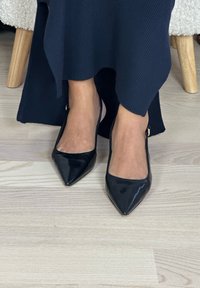 Femme portant des escarpins slingback noirs à bouts pointus et une longue jupe bleu marine foncé, assise sur un sol en bois clair près des pieds d'une chaise en bois.