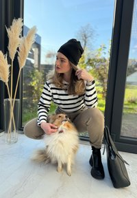 Μαύρο σκουφάκι, ριγέ μακρυμάνικο πουκάμισο και μπεζ παντελόνι. Γονατιστός δίπλα σε έναν αφράτο καστανό και λευκό σκύλο. Μαύρη τσάντα με υφασμάτινη λαβή.