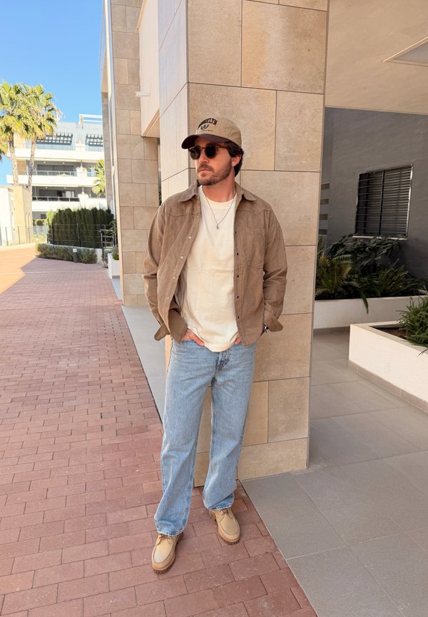 Mann mit beigefarbener Mütze, Sonnenbrille, brauner Jacke, weißem Hemd, hellblauen Jeans und beigen Schuhen, der draußen an einer gefliesten Gebäudeecke steht.