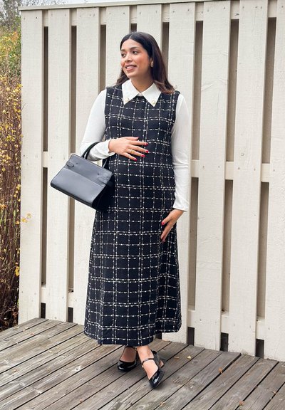 Vestido sin mangas con patrón de cuadrícula en blanco y negro, cuello blanco, combinado con un bolso negro y tacones con tira en el tobillo, sobre suelo de madera.