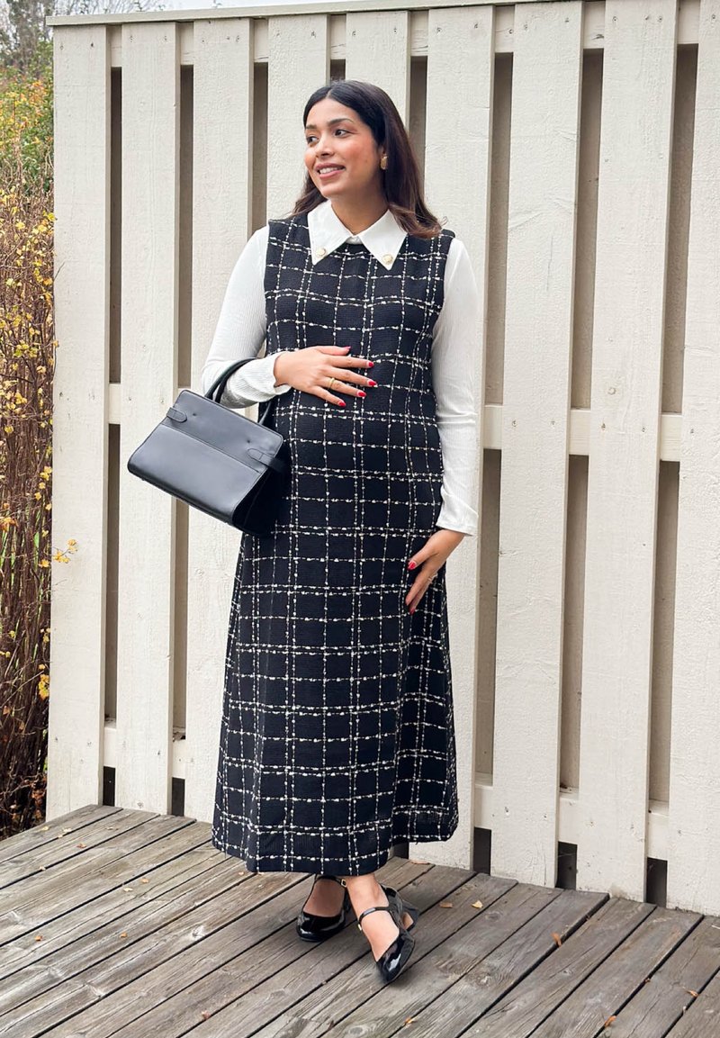 Robe sans manches à motif quadrillé noir et blanc, col blanc, associée à un sac à main noir et des escarpins à bride à la cheville, posée sur un sol en bois.