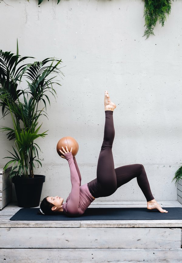 Figura femenina con una camiseta de manga larga color malva y leggings marrones realiza un ejercicio sobre una esterilla negra, sosteniendo una pelota de medicina color beige, rodeada de plantas.