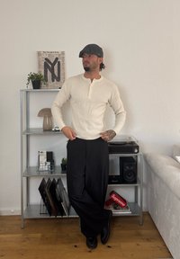 Beige ribbed long-sleeved henley shirt with button placket, teamed with black wide-leg trousers. A grey flat cap and black shoes complete the outfit.