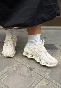 Os ténis Nike de cor clara apresentam um upper texturizado, acabamentos lisos e uma sola distintiva com amortecimento visível. Meias brancas com textura ribada completam o visual.