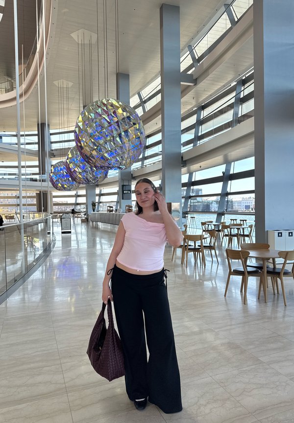 Femme en haut rose et pantalon noir tenant un grand sac, debout à l'intérieur d'un bâtiment moderne avec des lumières sphériques réfléchissantes et des murs en verre.