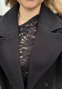 Abrigo negro de doble botonadura con solapas anchas sobre un top de encaje negro con motivos florales. Textura suave y diseño ajustado. Collar dorado visible.