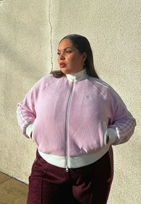 Veste zippée rose côtelée avec accents blancs et trois bandes sur les manches, accompagnée d'un pantalon bordeaux. Tissu lisse, coupe décontractée.