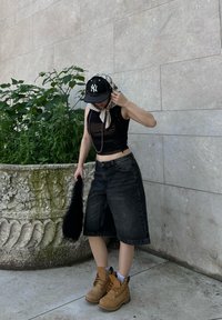 Črna skrajšana majica brez rokavov, preveliki temni jeans šortsi, svetlo rjavi čevlji na vezalke, črna kapa, svetla ruta, drži črn umetni krzneni dodatek.