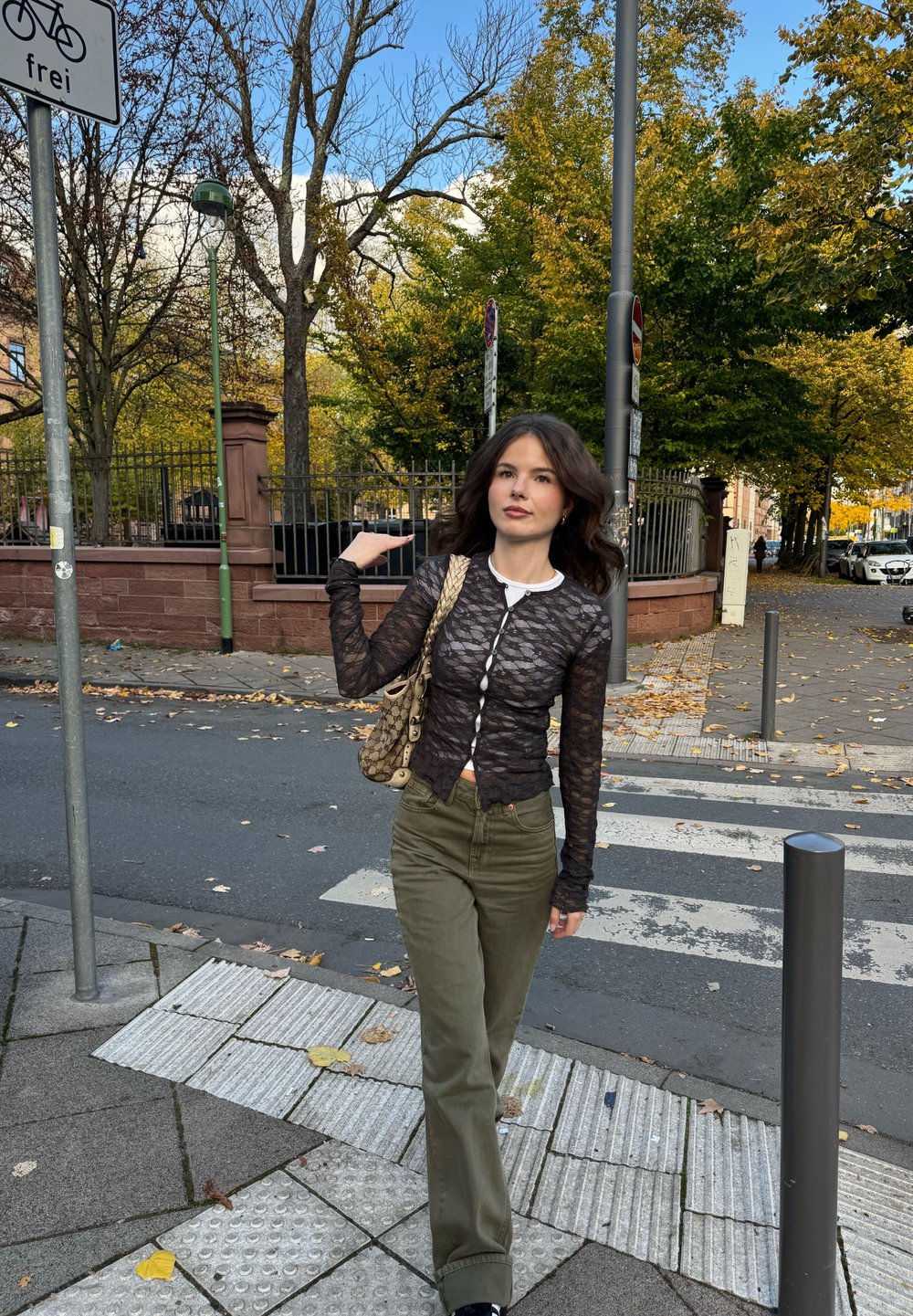 Crni dugih rukava čipkani top s gumbima, maslinasto zelene hlače visokog struka i torba s uzorkom preko ramena. Vanjski ambijent s drvećem i pločnikom.