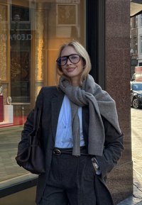 Grå uldfrakke over en hvid skjorte, kombineret med et oversized gråt tørklæde. Sorte briller med sort stel. Sort taske. Baggrunden viser et butiksvindue.
